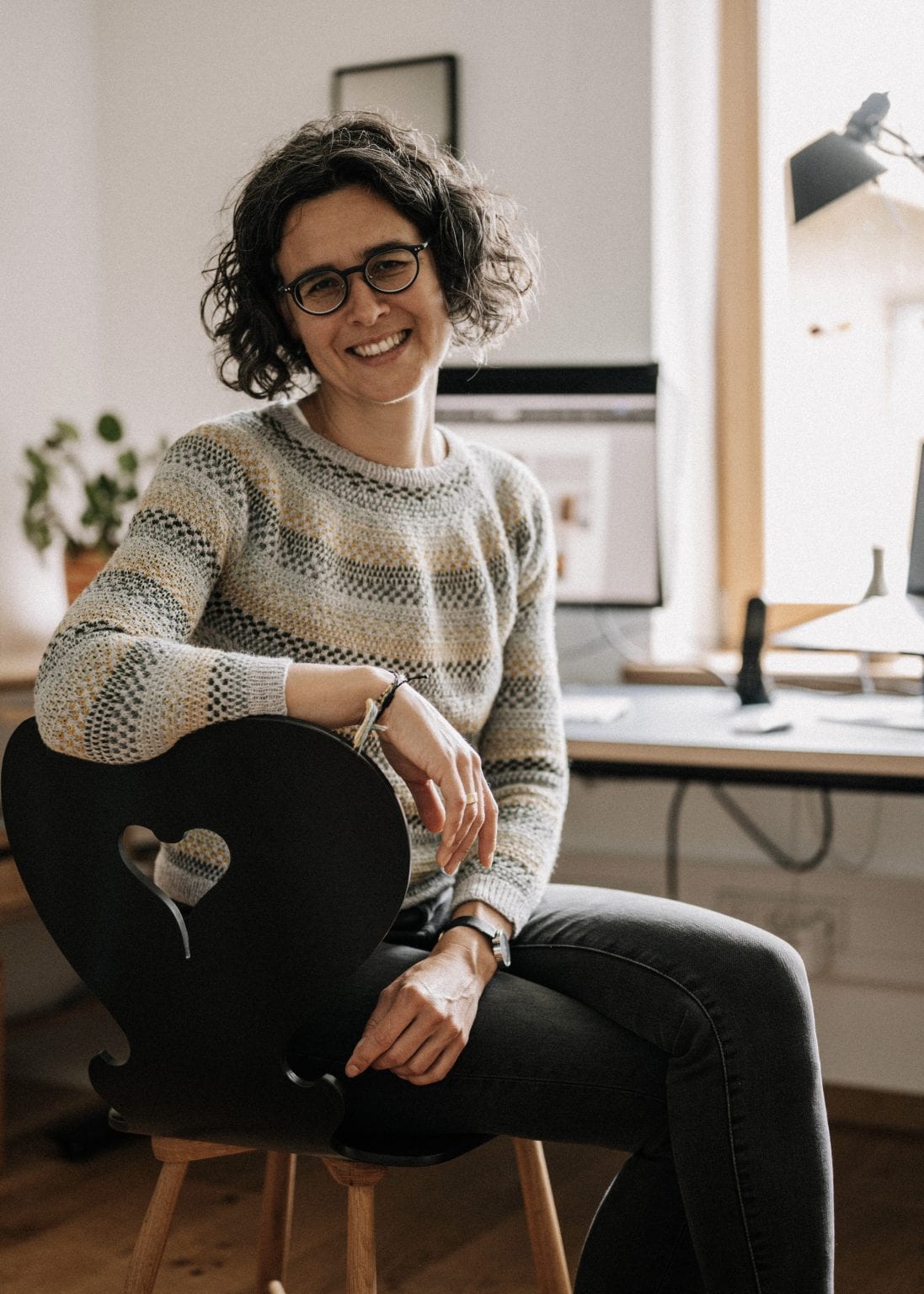 Barbara Prasch sitzt auf einem Holzstuhl und lächelt in die Kamera. Im Hintergrund sieht man ihren Schreibtisch mit Bildschirm.